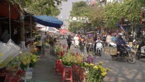 Static-shot-of-a-busy-street-with-moped-riders-passing-by-a-florist-in-Lang-Son