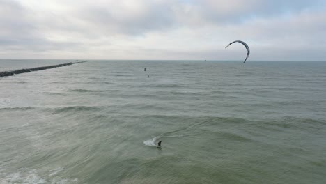 Establishing-aerial-view-of-a-group-of-people-engaged-in-kitesurfing,-overcast-winter-day,-high-waves,-extreme-sport,-Baltic-Sea-Karosta-beach-,-panoramic-drone-shot