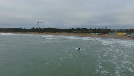 Establishing-aerial-view-of-a-man-engaged-in-kitesurfing,-overcast-winter-day,-high-waves,-extreme-sport,-Baltic-Sea-Karosta-beach-,-drone-shot-moving-forward