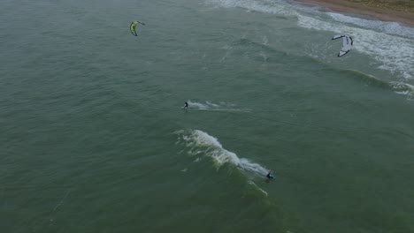 Establishing-aerial-view-of-a-group-of-people-engaged-in-kitesurfing,-overcast-winter-day,-high-waves,-extreme-sport,-Baltic-Sea-Karosta-beach-,-drone-birdseye-shot-moving-forward,-tilt-down