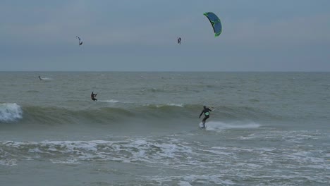 A-group-of-people-engaged-in-kitesurfing,-overcast-winter-day,-high-waves,-Baltic-Sea-Karosta-beach-,-slow-motion,-distant-shot