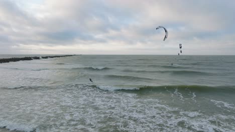 Establishing-aerial-view-of-a-group-of-people-engaged-in-kitesurfing,-overcast-winter-day,-high-waves,-extreme-sport,-big-jump,-Baltic-Sea-Karosta-beach-,-panoramic-drone-shot-moving-right