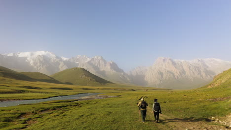 Two-hikers-on-trail-in-valley-with-the-Kakshaal-Too-mountain-range-in-the-distance,-Kyrgyzstan,-drone-flys-past-two-men