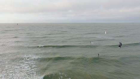 Establishing-aerial-view-of-a-group-of-people-engaged-in-kitesurfing,-overcast-winter-day,-high-waves,-extreme-sport,-Baltic-Sea-Karosta-beach-,-wide-drone-shot-moving-forward