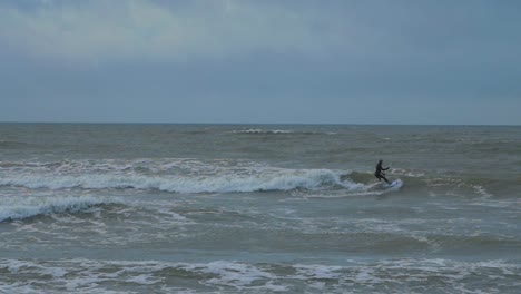 Man-engaged-in-kitesurfing,-overcast-winter-day,-high-waves,-Baltic-Sea-Karosta-beach-,-slow-motion,-distant-wide-shot