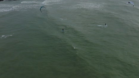 Establishing-aerial-birdseye-view-of-a-group-of-people-engaged-in-kitesurfing,-overcast-winter-day,-high-waves,-extreme-sport,-Baltic-Sea-Karosta-beach-,-drone-shot-moving-forward
