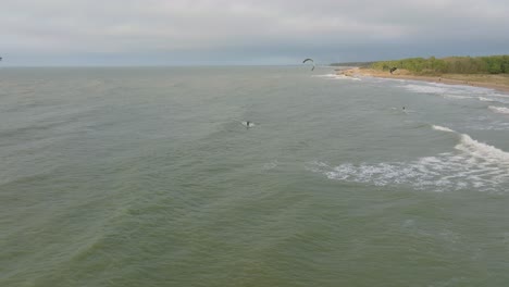 Establishing-aerial-view-of-a-group-of-people-engaged-in-kitesurfing,-overcast-winter-day,-high-waves,-extreme-sport,-Baltic-Sea-Karosta-beach-,-distant-drone-shot-moving-backward