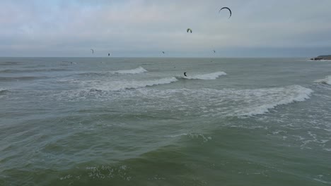 Establishing-aerial-view-of-a-group-of-people-engaged-in-kitesurfing,-overcast-winter-day,-high-waves,-extreme-sport,-Baltic-Sea-Karosta-beach-,-wide-drone-shot-moving-forward