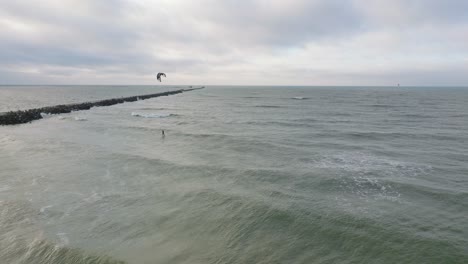 Establishing-aerial-view-of-a-man-engaged-in-kitesurfing,-overcast-winter-day,-high-waves,-extreme-sport,-Baltic-Sea-Karosta-beach-,-wide-drone-shot-moving-forward