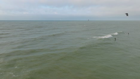 Establishing-aerial-view-of-a-group-of-people-engaged-in-kitesurfing,-overcast-winter-day,-high-waves,-extreme-sport,-Baltic-Sea-Karosta-beach-,-distant-wide-drone-shot-moving-forward