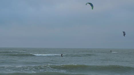 Eine-Gruppe-Von-Menschen,-Die-Kitesurfen,-Bewölkter-Wintertag,-Hohe-Wellen,-Ostsee-karosta-strand,-Zeitlupe,-Weitschuss
