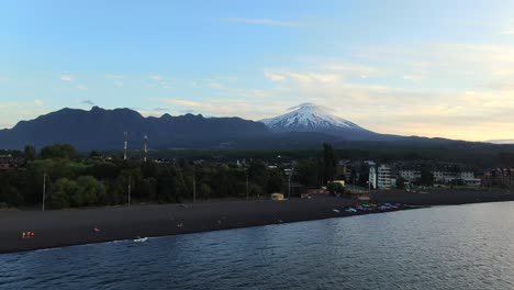 Aerial-approach-towards-shore-of-Villarrica-Lake-in-Pucon,-Chile-with-towering-Villarrica-Volcano-in-background