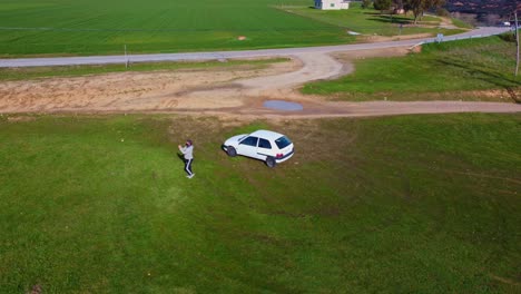 Luftbild-Mann,-Der-Eine-Drohne-In-Der-Nähe-Eines-Kleinen-Autos-Steuert,-Umgeben-Von-Grünen-Wiesen-Und-Landschaft