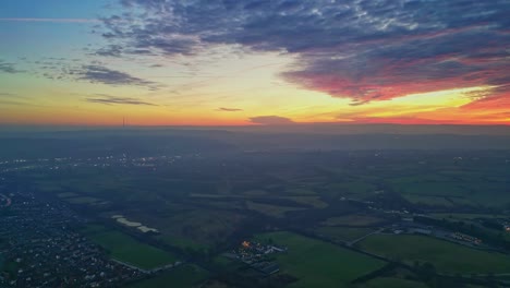Drone-Cinematic-Town-and-Countryside-Scenery-Green-Misty-Fields-Colorful-Red-Sunset-Horizon-Fiery-Clouds