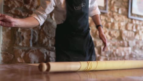 Heldenaufnahme-Eines-Italienischen-Kochs,-Der-Mehl-Auf-Einen-Holztisch-Zu-Einem-Handgemachten-Stück-Pasta-Mit-Steinmauerhintergrund-Wirft