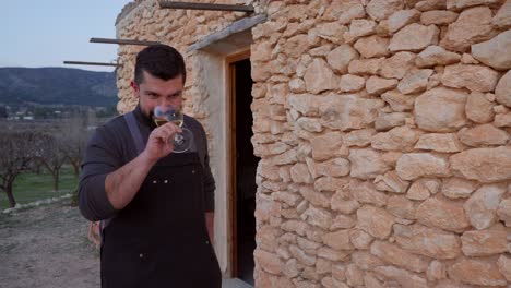 A-smiling-bearded-man-toasts-with-a-glass-of-white-wine-in-the-background-of-a-typical-Mediterranean-stone-hut