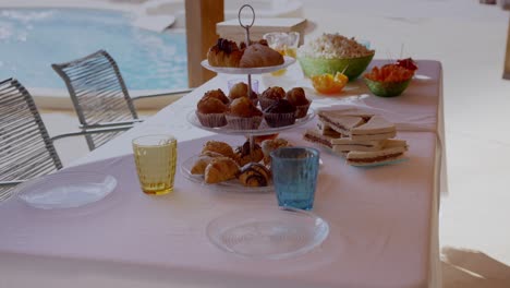 Table-with-sweets-for-a-poolside-party-in-summer,-with-a-tray-with-croissants,-sandwich,-popcorn,-crudités,-coloured-glasses,-transparent-plates-and-white-tablecloth
