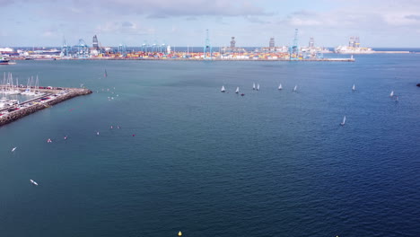 Aerial-shot-over-the-water,-sailing-boats-and-surf-ski-boats,-pier-on-the-left-and-port-on-the-horizon