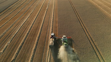 Falling-Establishing-Drone-Shot-of-Back-of-Claas-Combine-Harvester-with-Arm-Out-Unloading-Grain-into-Tractor-with-Trailer-Full-of-Grain-and-then-Pulls-Away-at-Golden-Hour-Sunset-UK