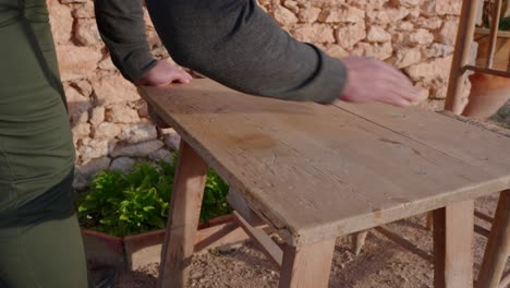 A-man-preparing-a-wood-table-for-a-meal,-in-the-early-spring-in-the-countryside-in-an-old-stone-house-among-Mediterranean-olive-trees
