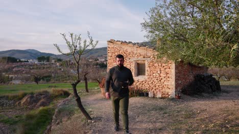 Man-walks-among-almond-blossom-trees-at-sunset,-with-an-old-Mediterranean-style-stone-hut-in-early-spring