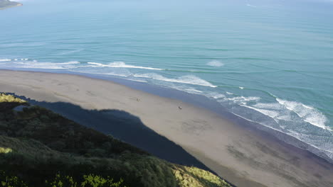 A-pair-of-people-ride-Horseback-at-a-beach-in-Raglan,-New-Zealand-along-hillsides-and-mountains-taken-by-drone