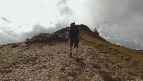 Third-person-view-of-a-man-hiking-on-a-rocky-path-towards-a-mountain-peak-with-storm-clouds-on-the-horizon