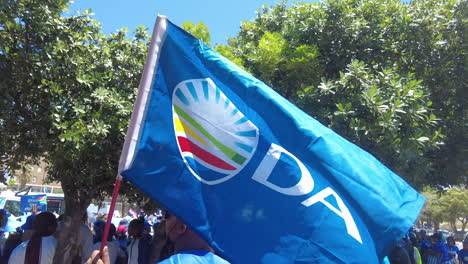 Una-Bandera-Del-Partido-De-La-Alianza-Democrática-Ondeando-En-El-Viento-Mientras-Los-Manifestantes-Se-Congregan-Para-Marchar-Contra-Los-Cortes-De-Carga-Y-Los-Apagones-Continuos-De-Eskom