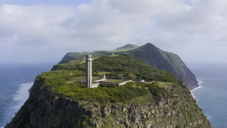 Pull-back-drone-footage-of-a-Lighthouse-revealing-dramatic-cliffs-and-the-Atlantic-Ocean-and-the-São-Jorge-island-shape,-in-the-Azores,-Portugal