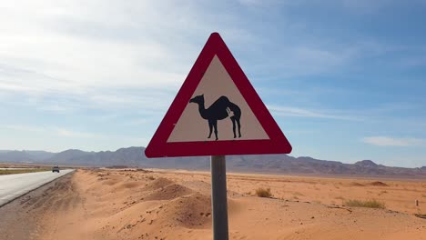Triangle-road-sign-with-camel-warning-located-in-the-wilderness-of-Arabian-desert-with-red-sand-and-rugged-mountains-landscape-with-blue-sky-in-Jordan,-Middle-East