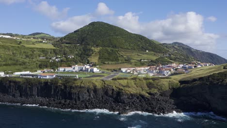 Velas,-Sao-Jorge-island,-Azores,-Portugal-Aerial-view-of-whitewashed-houses-village,-skate-park,-sports-field,-with-surrounding-mountains,-vegetation-and-lava-formations-seaside-drone-footage