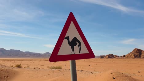 Triangle-road-sign-with-camel-in-the-vast-remoteness-of-desert-landscape-with-red-sand-terrain-and-mountains-in-Jordan,-Middle-East