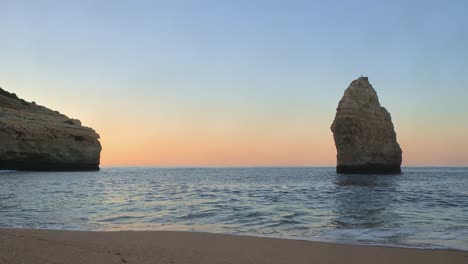 Ground-level-footage-of-gentle-waves-lapping-on-a-beautiful-deserted-beach-in-Algarve,-Portugal-at-sunrise-showing-sandstone-cliffs-and-a-sea-stack