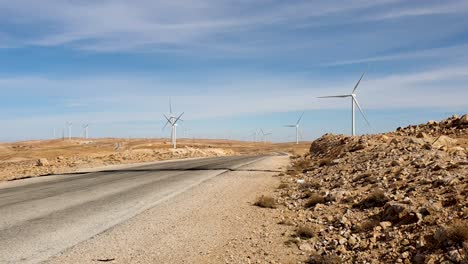 Car-driving-on-long,-straight-road-through-a-wind-turbine-farm-in-remote-desert-landscape-of-Jordan,-Middle-East