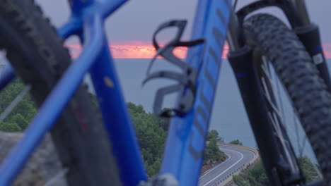 Time-lapse-of-mountain-bike-frame-in-front-of-a-road-bend-as-cars-drive-past-while-sun-rises-over-the-ocean-in-the-background