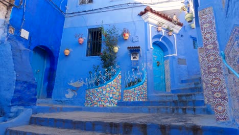 Vista-Inclinada-Hacia-Abajo-A-Través-De-Una-Hermosa-Puerta-Azul-Con-Dos-Pequeños-Jardines-De-Flores-Al-Lado-De-Una-Calle-Angosta-Con-Escalones-En-Chefchaouen-En-Marruecos