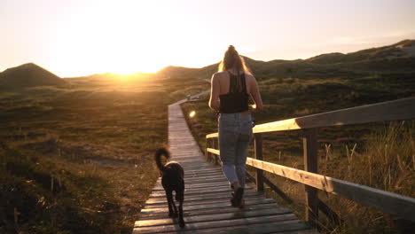 Woman-and-her-dog-walking-on-a-wooden-boardwalk-while-sunset-in-a-dune