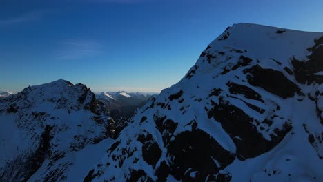 Aerial-trucking-shot-of-beautiful-snowy-mountain-range-against-blue-sky-and-sunlight-at-sunrise---Jonshornet,-Norway