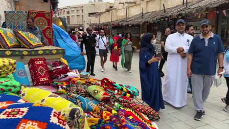 Bunte-Handgefertigte-Gewebte-Kissen-Und-Textilhandwerk-Auf-Einem-Traditionellen-Lokalen-Markt-In-Doha-Katar-Msheireb-Altstadt-Innenstadt-Menschen-Mit-Hijab-Islamischer-Kleidung-Für-Frauen-Und-Arabischer-Kleidung-Für-Männer