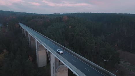 Aerial-View-of-White-Electric-Car-Moving-on-Empty-Bridge-Road-in-Twilight,-Tracking-Drone-Shot