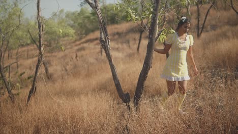 Serene-footage-of-a-young-beautiful-Indian-girl-walking-coyly-around-a-tree-on-a-hill-in-a-field-of-tall-dead-yellow-grass