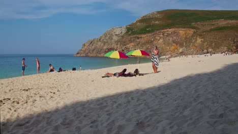 People-Enjoying-The-Waters-Along-The-Shore-And-Some-Relaxing-Under-Umbrella-At-Porth-Beach-In-Cornwall,-England,-UK