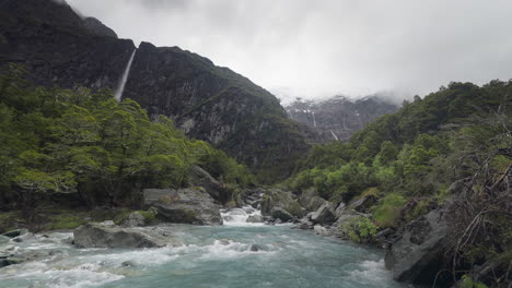 Glacial-river-rapids-in-lush-green-valley-in-New-Zealand-with-waterfalls-in-Background