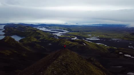 Aerial-drone-landscape-view-of-one-person-wearing-a-red-jacket,-standing-on-top-of-a-mountain,-in-the-Iceland-highlands