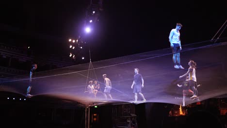 Cirque-Du-Soleil:-Atletas-De-Circo-Profesionales-Practicando-Y-Ensayando-El-Acto-De-Trampolín-En-Un-Impresionante-Lugar-Con-Luz-Tenue,-Hombre-Cayendo-Del-Trapecio