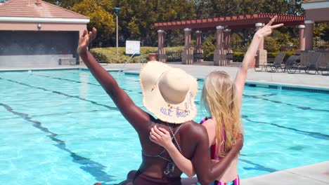 Friends-at-the-pool,-sitting-on-the-edge-and-waving-with-their-hands,-interracial-friendship-concept