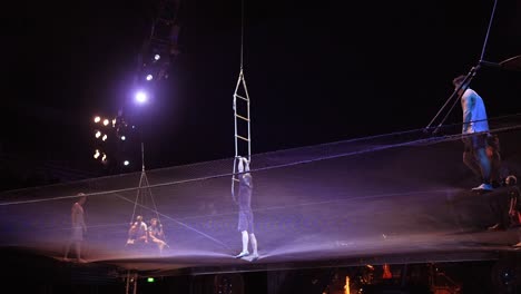 Cirque-Du-Soleil:-Atletas-De-Circo-Profesionales-Practicando-Y-Ensayando-El-Acto-De-Trampolín-En-Un-Impresionante-Lugar-Con-Poca-Luz,-Hombre-Cayendo-Del-Trapecio,-Hombre-Levantado-Por-Una-Escalera-De-Cuerda