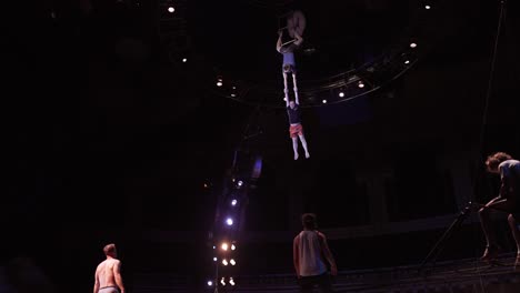 Cirque-Du-Soleil:-Professionelle-Zirkussportler-üben-Und-Proben-Trampolin-Darbietungen-In-Einem-Atemberaubenden-Veranstaltungsort-Mit-Gedämpftem-Licht,-Mann-Fällt-Vom-Trapez-Und-Macht-Rückwärtssaltos