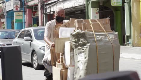 Ein-älterer-Mann-Mit-Maske,-Der-Einen-Mit-Pappe-Aus-Müll-Gefüllten-Karren-Entlang-Der-Belebten-Straße-Von-Kuala-Lumpur-Schiebt