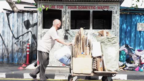 Alter-Asiatischer-Mann-Schiebt-Wagen-Mit-Gesammeltem-Karton,-Müllcontainer-Im-Hintergrund